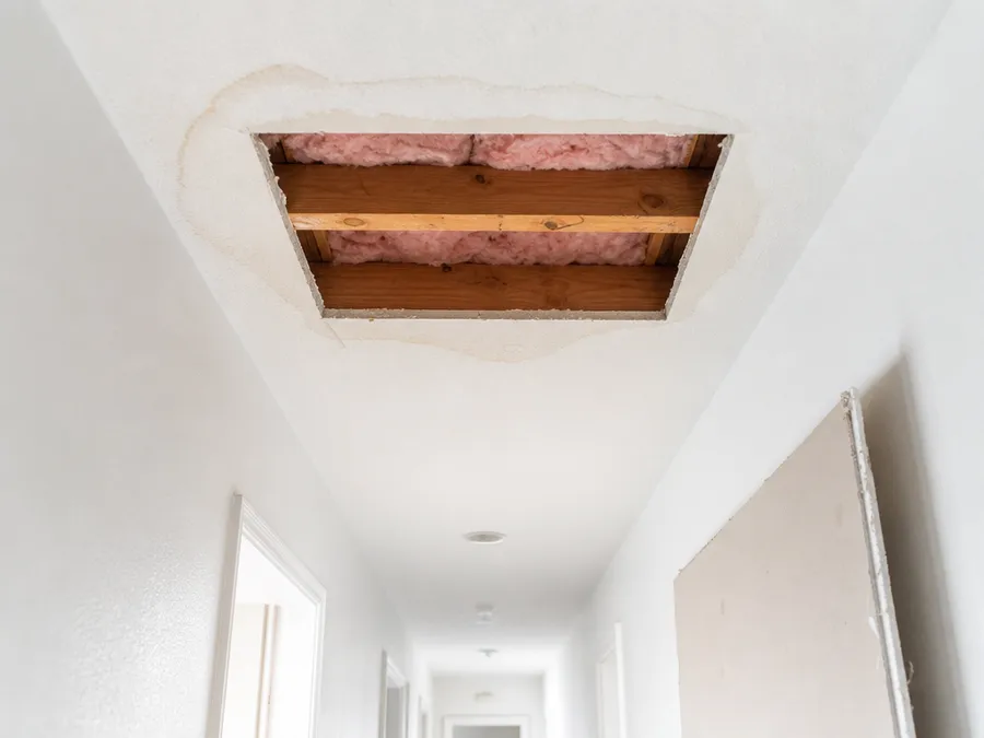 A hallway ceiling with a rectangle of water-damaged drywall cut out, showing the joists and insulation above, and a new drywall sheet leaning below