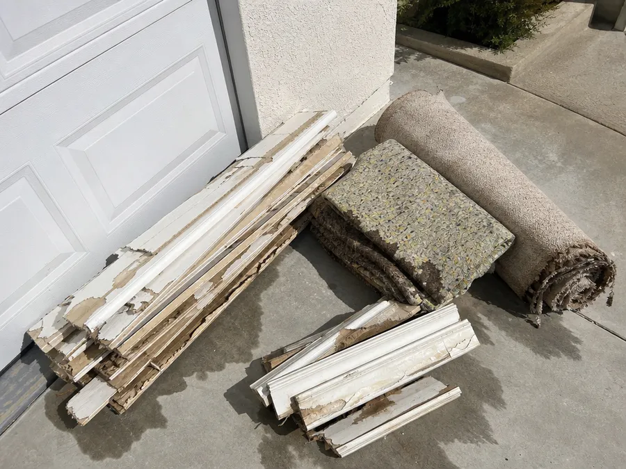 Water-damaged drywall strips, rolled carpet, carpet pad and baseboard stacked on a driveway after removal