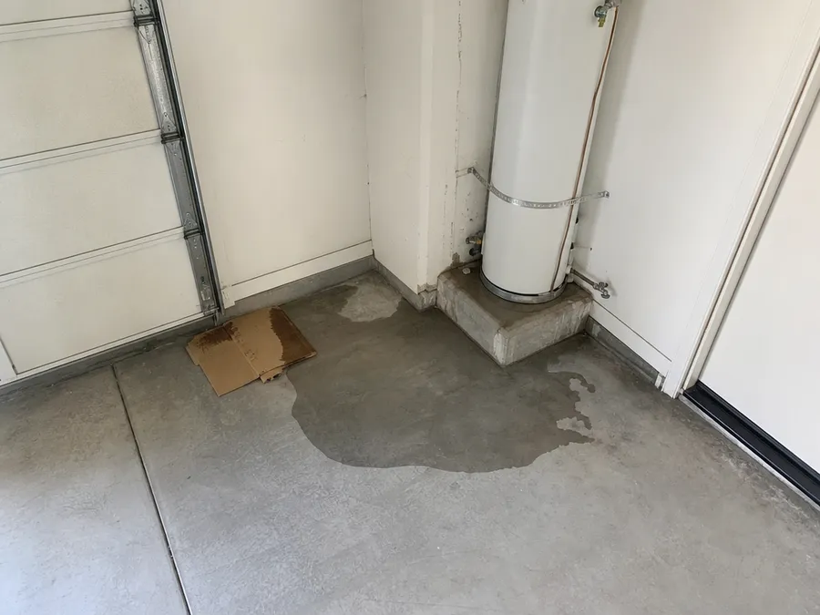 A tank water heater on a raised platform in a garage corner, with water spreading across the concrete floor and into the base of the wall