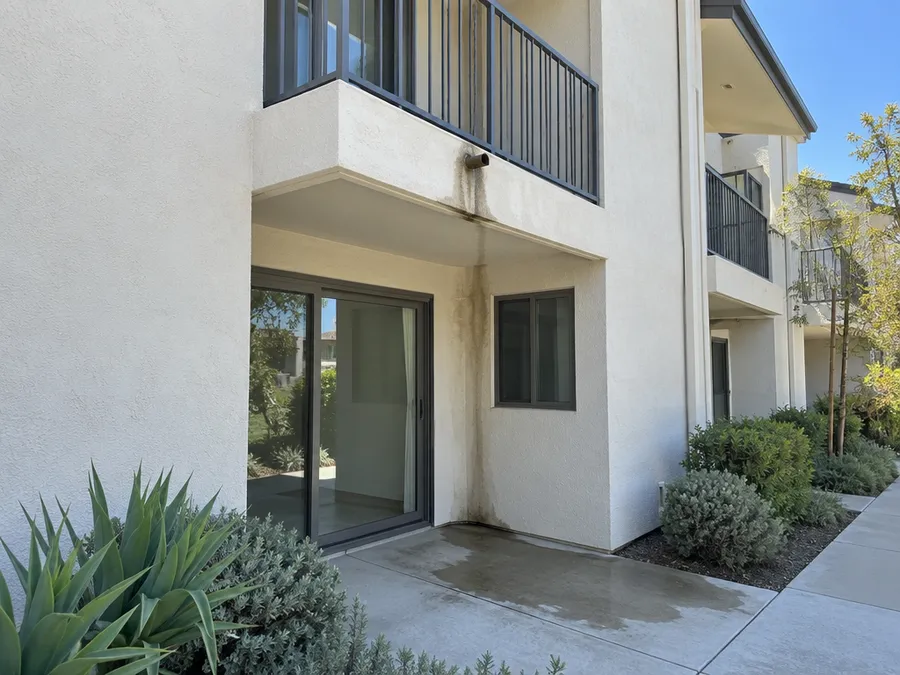 A dark water streak running down a pale stucco wall from a small balcony drain spout to a damp patio and sliding door below