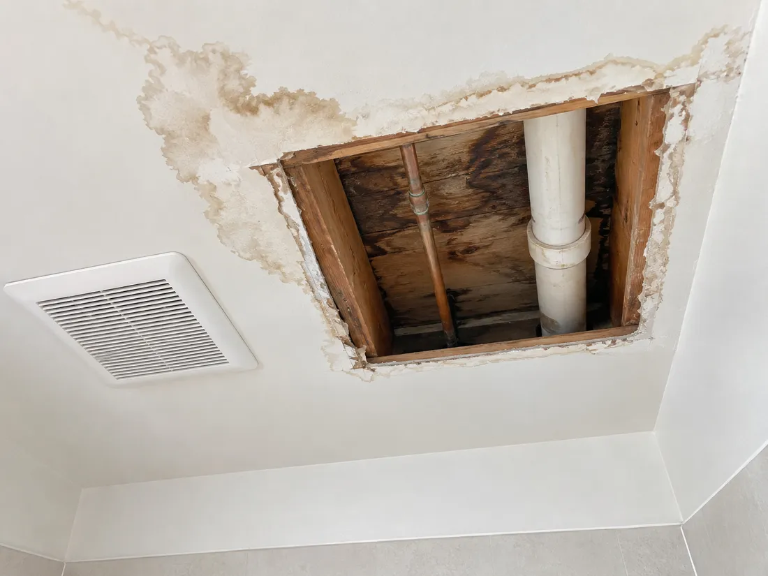 A bathroom ceiling with a square of drywall cut away, showing a copper supply pipe, a white drain pipe and water-stained joists above