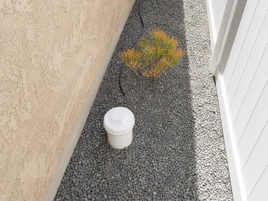 A white sewer cleanout pipe with a threaded cap in a gravel side yard beside a stucco wall