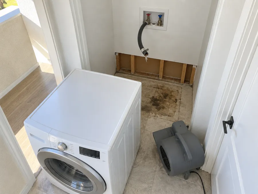 A washing machine pulled out of its alcove beside a split fill hose, the lower drywall behind it cut away to the studs