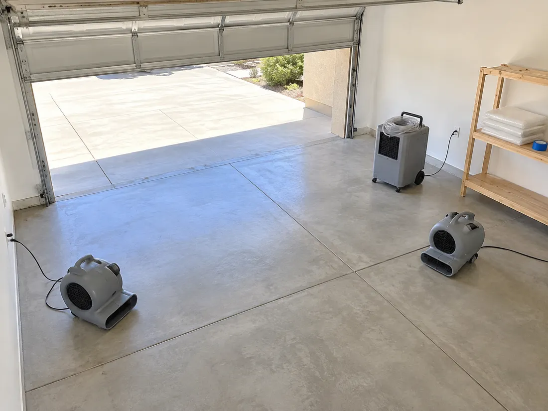 Two grey air movers and a dehumidifier standing on a clean concrete garage floor, the door open to daylight